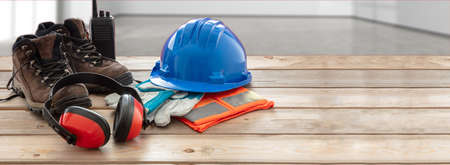Work safety protection equipment. Industrial protective gear on wooden table, blur office or home interior background. Hardhat, boots, earmuffs, gloves, gogglesの写真素材