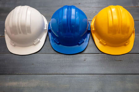 Work safety protection equipment. Industrial protective hardhats white, yellow and blue colors on wooden background. Personal health and safety concept.の写真素材