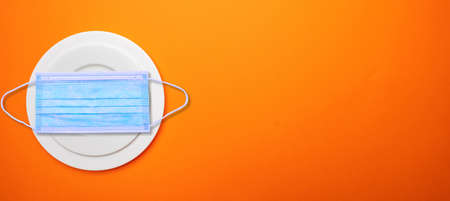Coronavirus for food and restaurant concept. Face protective mask on a plate set against orange color background, copy space. COVID spread prevention measureの写真素材