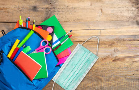 School, coronavirus days. School supplies and medical protective face mask out of a backbag, wooden desk  background, top view, copy spaceの写真素材