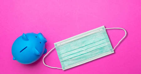 Money saving in coronavirus pandemic concept. Protective face mask and blue piggy bank, pink color background, copy space. COVID19 affects economy,の写真素材