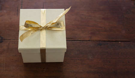 Surprise with present concept. Beige closed gift box packaging with gold color ribbon against brown wooden table background. Copyspace, high angle view.の写真素材