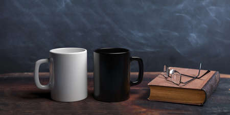 Black and white coffee mugs, old books and reading glasses on a wooden table, chalkboard background. 3d illustrationの写真素材