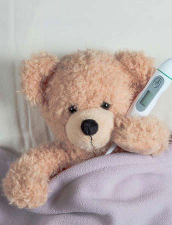 Fever, kids cold, covid virus flu concept. Cute teddy with a thermometer in bed, covered with a warm blanketの写真素材