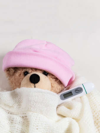 Fever, kids cold, covid virus flu concept. Cute teddy with a thermometer in bed, covered with a warm blanketの写真素材