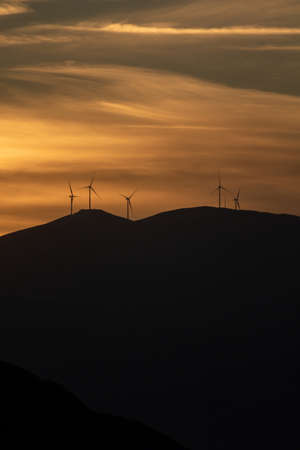 Sunrise, sunset on colorful sky. Golden sunbeams paint the horizon, wind generators on hills give energy power. Sun through clouds over mountains silhouette, cloudscape background. Vertical photo.の写真素材
