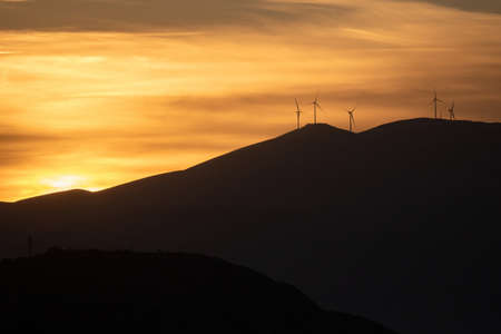 Sunrise, sunset on colorful sky. Golden sunbeams lighten the horizon, wind generators on hills give electricity. Sun through clouds over mountains silhouette, cloudscape background. Wallpaper, card.の写真素材