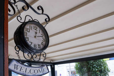 Welcome sign, vintage clock. Low angle view of shabby retro clock hanging under roof on porch...