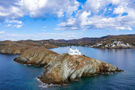 Greece, Kea Tzia island. Aerial drone view of lighthouse and white St. Nicolas church on a rocky cape, cloudy blue sky and transparent rippled sea water background. Summer day, tourist destination.の写真素材