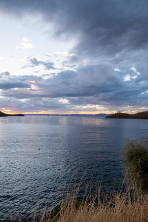 Greece, Kea Tzia island. White lighthouse on a rocky cape, cloudy blue sky, wild dry plants and rippled sea sparkle water background. Summer, afternoon, tourist destination. Vertical view.の写真素材
