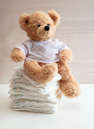 Baby diapers stack, cute teddy bear sitting on top, white color floor, wooden wall background. Newborn nappies conceptの写真素材