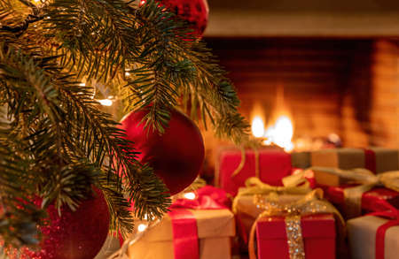 Xmas home celebration concept. Christmas tree branch with red ornament balls, gift boxes with ribbons, bows and blur burning fireplace background. Warm family moments.の写真素材
