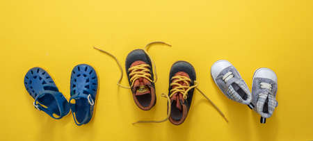 Baby first steps. Three pairs of boy blue shoes variety on yellow color background, top view. Kids footwear, beach sandals, booties and soft infant shoes in a row.の写真素材