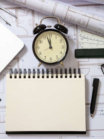Construction design deadline. Engineer blank notebook and alarm clock on project blueprints...