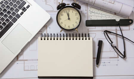 Construction design deadline. Architect engineer blank notebook, laptop and alarm clock on project blueprints background.の写真素材