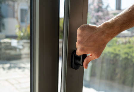 Tilt and turn grey color aluminum window, man holding the handle, fresh air for home. Male hand vertical open metal or PVC window, closeup view.の写真素材