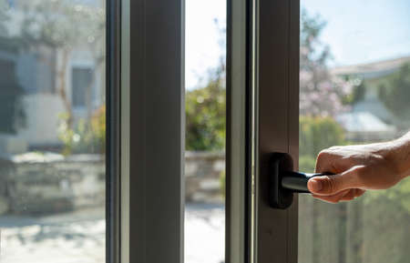 Hand open aluminum window frame closeup view. Man holding the metal or PVC door handle. Energy efficient, security profile, blur garden outdoor backgroundの写真素材