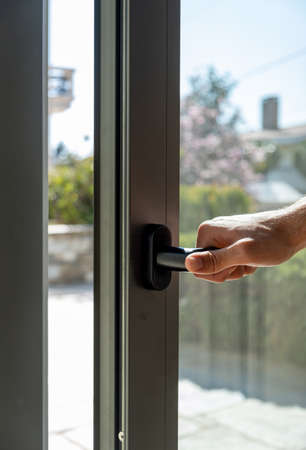 Male hand open aluminum window frame closeup view. Man holding the metal or PVC door handle. Energy efficient, security profile, blur garden outdoor backgroundの写真素材