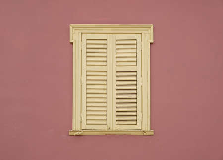 Window wooden shutters on brown color painted wall background. Yellow pale french style closed window blinds with ornamental frame on residential building facadeの写真素材