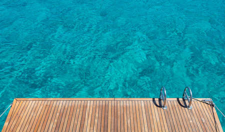 Yacht stern deck, teak wood and metal ladders, turquoise blue sea water background. Sailing boat...