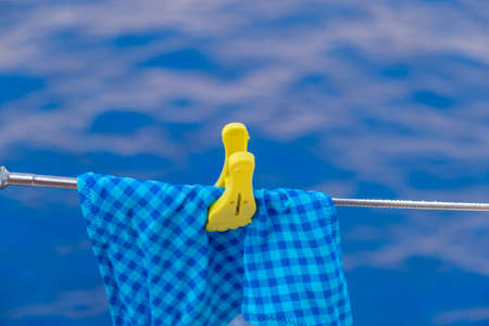 Swimwear plaid blue hanging with yellow pin on boat stainless steel rail. Wet swimsuit drying on clothesline, sea water background. Summer holidays and sailing. Copy space, advertise templateの写真素材
