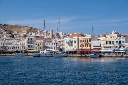 Syros island, Cyclades, Greece. May 26, 2021. Destination Hermoupolis capital of Siros or Syra town background. Port with moored yachts, waterfront cafe, shops, whitewashed buildings sea sun blue sky.のeditorial素材