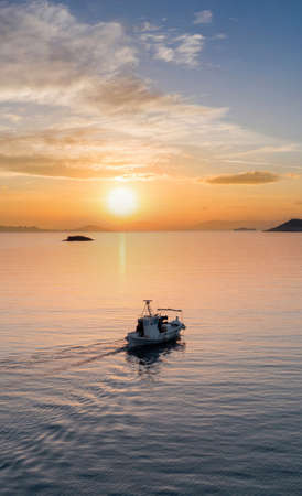 Fishing boat at sunset. Cyclades island. Greece. Aerial drone view of an outbound typical wooden fishing boat moving on calm sea and orange color sky background.の写真素材