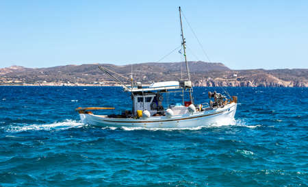 Cyclades, Greece. Fishing boat white traditional wooden vessel sailing in rough wavy sea. Rocky land, Greek island coast and clear blue sky background.のeditorial素材