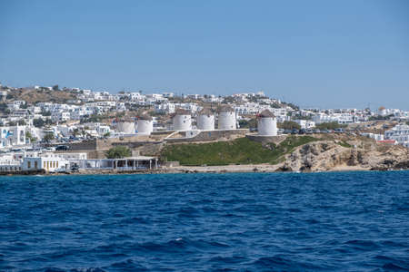 Mykonos, Cyclades, Greece. Traditional windmills  famous landmark and cityscape. Summer cosmopolitan destination for vacation, taverns cafe on the seaside, clear blue sky background. View from the seaの写真素材