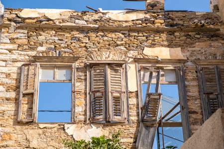 Damaged stone wall building broken aged wooden shutters and windows with blue sky view. Abandoned...
