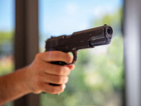 Pistol in male hand. Man aiming with a gun, blur glass window background, closeup view. Killer,...