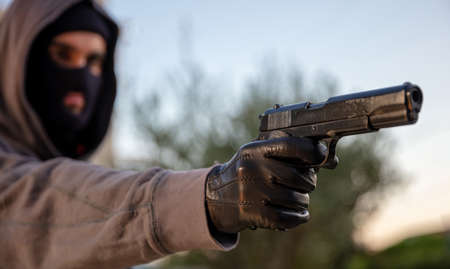Man aiming with a pistol, nature outdoors background, closeup view. Killer hooded with balaclava,...