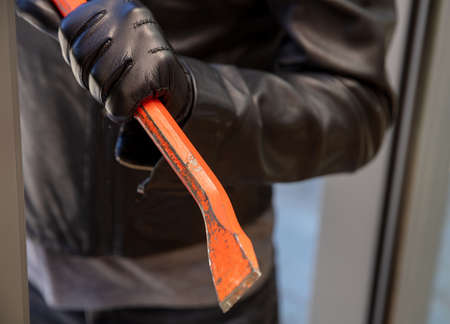 Masked thief entering a house for stealing in the daytime. Burglar in leather jacket breaking a glass aluminum door. Housebreak with crowbar.の写真素材