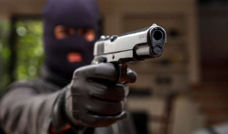 Give me your money otherwise you are dead. Blur robber with black balaclava aiming with pistol the...