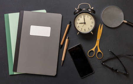 School teacher or student office desk flat lay, workspace. Notebooks, mobile phone stationery and alarm clock on black color background, top view.の写真素材