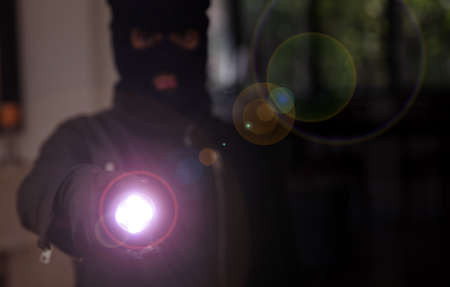 Housebreaking, burglary concept. Masked burglar in black balaclava entering a house holding a flashlight, dark night background.の写真素材