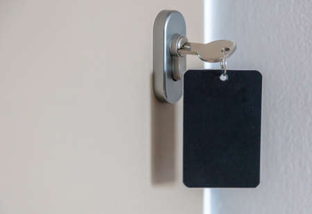 Door key with blank label in the keyhole. Steel key ring, room key and empty black tag for text or number close up view. House, hotel or motel room open, unlock concept. Copy space, templateの写真素材