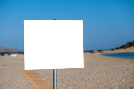 Sign white blank on post, sandy beach background. Outdoors empty signboard billboard signage label on metal pole. Sunny day summer holiday advertise template, copy space.の写真素材
