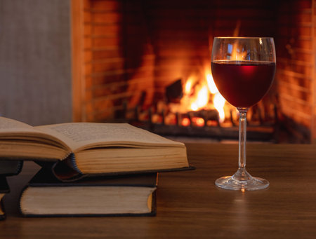 Red wine drinking and reading a book by the fire. A wineglass and an open book against burning fireplace background. Winter holidays relaxation, cozy warm home interiorの写真素材