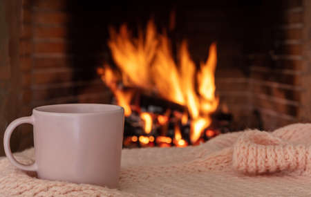 Cup of coffee on a sofa blanket, burning fireplace background. Pink color tea mug, relaxation by the fire, Christmas holiday cozy warm homeの写真素材