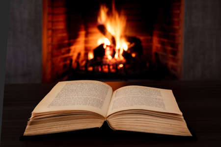 Reading and relaxation near the fire, Book open, fireplace burning firewood background. Christmas holiday cozy warm home interior.の写真素材