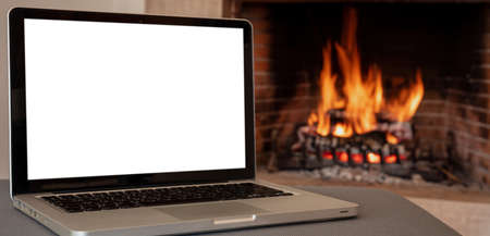 Winter home office concept. Blank screen laptop on burning fireplace background, White empty computer display, cozy room interior. Christmas warm houseの写真素材