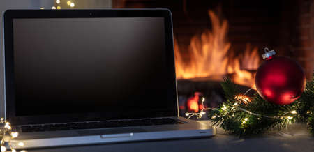 Christmas home office concept. Blank screen laptop and Xmas decoration on burning fireplace background, Winter holidays cozy warm house, space, templateの写真素材