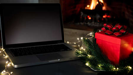 Blank screen laptop and Christmas present on burning fireplace background, Winter holidays home office concept. Xmas cozy warm houseの写真素材