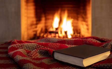 Book on a blanket, fireplace burning firewood background. Reading and relax near the fire, Christmas holiday cozy warm home interior.の写真素材