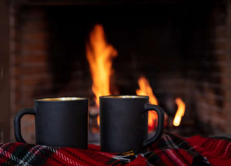 Coffee cups on a sofa red plaid blanket, burning fireplace background. Christmas holiday cozy warm...