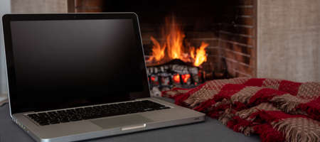 Winter home office concept. Blank screen laptop on burning fireplace background, Black empty computer display, cozy room interior. Christmas warm houseの写真素材
