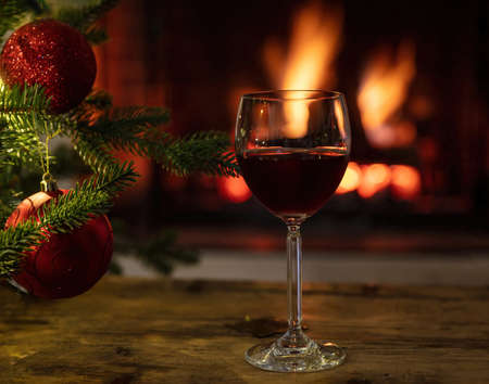 Christmas, red wine glass on a wooden table, burning fireplace background. Xmas tree decoration. Cozy warm home interior, winter holidays relaxationの写真素材