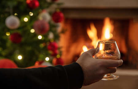 Man holding a brandy glass, Christmas tree and burning fireplace background. Drink alcohol and relax, winter holidays. Warm cozy home interiorの写真素材