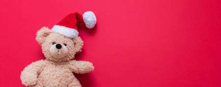Christmas present. Teddy bear wearing Santa hat on red background, banner, copy space. Kids holidays gifts, toys and stuffed animals. Holiday greeting card.の写真素材